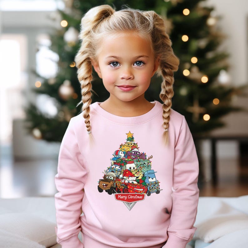 May include: A light pink sweatshirt featuring a Christmas tree design made of cartoon cars. The cars are in various colors, and the tree is topped with a gold star. The words "Merry Christmas" are printed at the base of the tree.