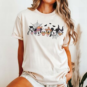 Puede incluir: Una camiseta de color crema con un gr&aacute;fico de Mickey Mouse, Minnie Mouse, Goofy, Donald Duck, Pluto y Mal&eacute;fica. El gr&aacute;fico presenta una telara&ntilde;a y murci&eacute;lagos.