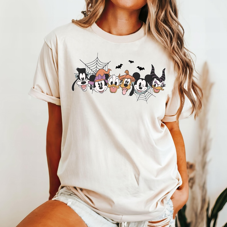 Puede incluir: Una camiseta de color crema con un gr&aacute;fico de Mickey Mouse, Minnie Mouse, Goofy, Donald Duck, Pluto y Mal&eacute;fica. El gr&aacute;fico presenta una telara&ntilde;a y murci&eacute;lagos.
