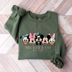 May include: Green sweatshirt with a graphic of Mickey Mouse, Minnie Mouse, Donald Duck, Goofy, and Pluto. The text "MICKEY & CO. Est. 1928" is printed below the graphic.