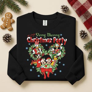 Op de afbeelding: Zwarte sweatshirt met een kerstontwerp met Mickey Mouse en vrienden. De tekst "Mickey's Very Merry Christmas Party" staat boven de krans. Het ontwerp bevat rood, groen en wit.