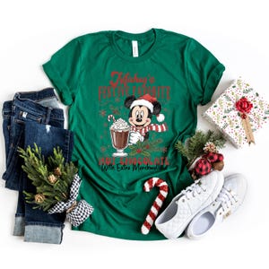 May include: A green t-shirt with the text "Mickey's Festive Favorite Hot Chocolate With Extra Marshmallows" featuring Mickey Mouse in a Santa hat and scarf, holding a hot chocolate. The image includes blue jeans, white shoes, and Christmas decorations.