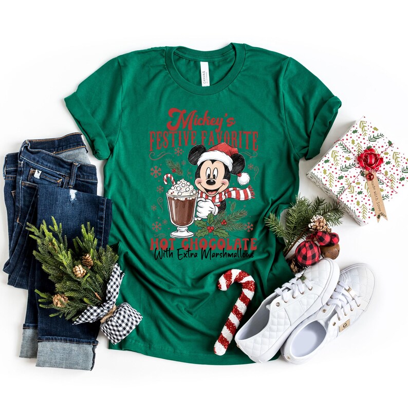 May include: A green t-shirt with the text "Mickey's Festive Favorite Hot Chocolate With Extra Marshmallows" featuring Mickey Mouse in a Santa hat and scarf, holding a hot chocolate. The image includes blue jeans, white shoes, and Christmas decorations.