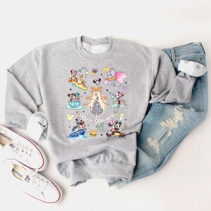 May include: A gray sweatshirt with a colorful print of various Disney characters, including Mickey Mouse, Minnie Mouse, Donald Duck, and Goofy. The print also includes the iconic Cinderella's Castle and the text "Best Day Ever".