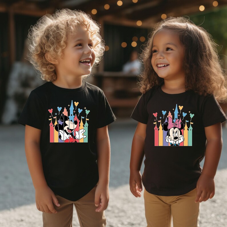 May include: Two children wearing black t-shirts with colorful graphic prints of Mickey Mouse and Minnie Mouse in front of a castle. The shirts feature a rainbow of colors and the text "Disney".