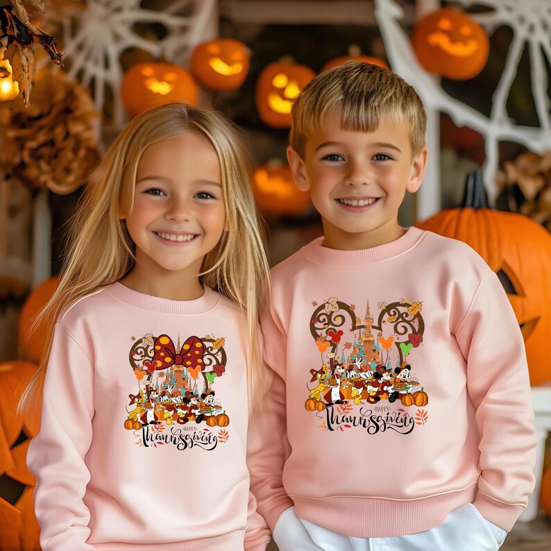 May include: Two children wearing matching light pink sweatshirts with a Disney-themed Thanksgiving design. The design features Mickey and Minnie Mouse, pumpkins, and the text "Happy Thanksgiving". The background includes carved pumpkins.