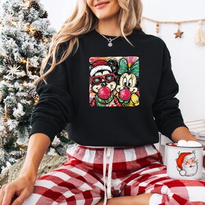 May include: Black sweatshirt featuring a colorful graphic of Mickey and Minnie Mouse blowing bubblegum bubbles. Mickey wears a Santa hat and sunglasses. Minnie has a green bow. The sweatshirt is paired with red and white plaid pajama pants.
