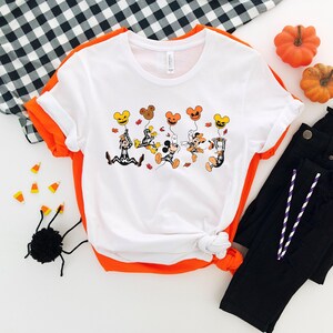 May include: White t-shirt with orange trim featuring a Halloween cartoon design. The graphic shows cartoon characters holding pumpkin-shaped balloons. The shirt is paired with black trousers and Halloween decorations.