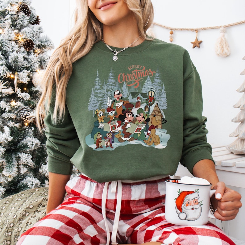 May include: Green sweatshirt with a graphic of Disney characters singing Christmas carols. The words "Merry Christmas" are above the characters. The person is wearing red and white plaid pajama pants and holding a mug with a Santa Claus design.