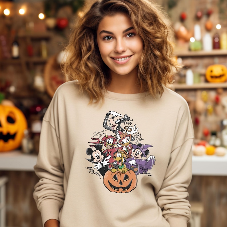 May include: Beige sweatshirt featuring a Halloween-themed graphic. The graphic includes cartoon characters like Mickey Mouse, Donald Duck, and Minnie Mouse, along with a carved pumpkin. The design is in shades of black, red, orange, and purple.