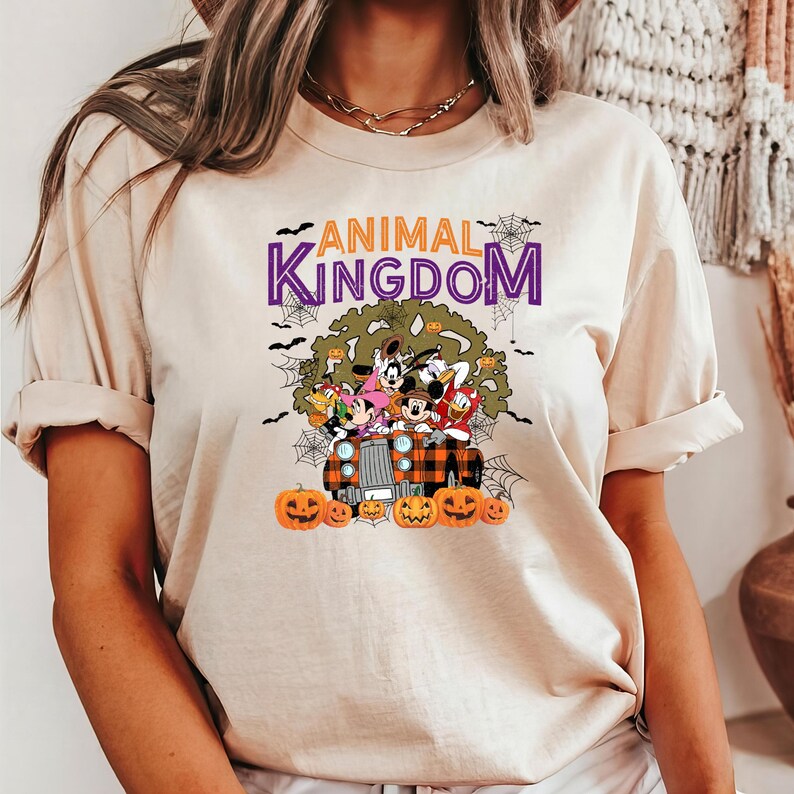 May include: Beige t-shirt with a Halloween design. The graphic includes the text "ANIMAL KINGDOM" in purple, cartoon characters, pumpkins, and spiderwebs. The characters are riding in a plaid truck.