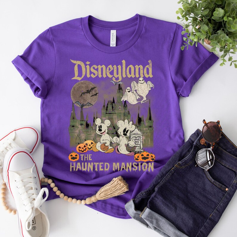 May include: Purple t-shirt with a vintage-style graphic of Disneyland's Haunted Mansion. The graphic features Mickey Mouse and a ghost in front of the Haunted Mansion, with a moon and bats in the background. The text "Disneyland" and "The Haunted Mansion" are also included in the graphic.