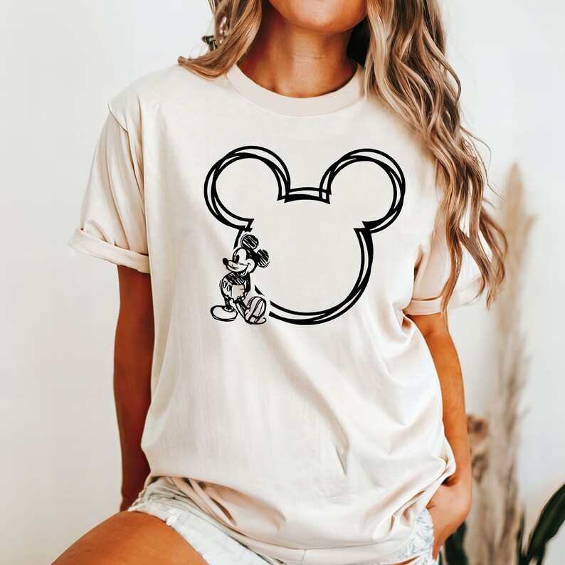 May include: A cream-colored t-shirt with a black outline of Mickey Mouse's head and a sketch of Mickey Mouse standing to the left of the outline.