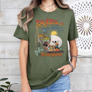 May include: A green t-shirt with a vintage-style graphic of Mickey Mouse and other Disney characters on a pirate ship. The text "Pirates of the Caribbean Disneyland" is printed on the shirt.