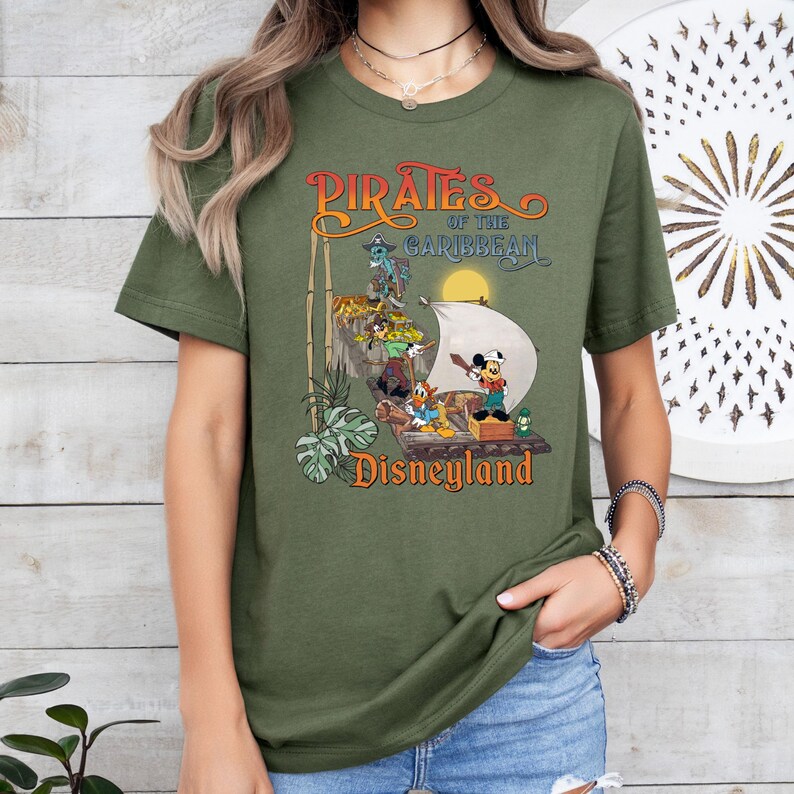 May include: A green t-shirt with a vintage-style graphic of Mickey Mouse and other Disney characters on a pirate ship. The text "Pirates of the Caribbean Disneyland" is printed on the shirt.