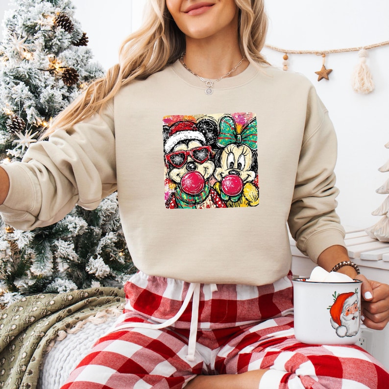 May include: Beige sweatshirt featuring a colorful graphic of Mickey and Minnie Mouse blowing bubblegum. Mickey wears sunglasses and a Santa hat. Minnie has a green bow. The sweatshirt is paired with red and white plaid pajama pants.