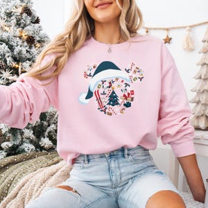 May include: A light pink sweatshirt featuring a Christmas-themed Mickey Mouse design. The design includes a Santa hat, a Christmas tree, and various holiday elements. The sweatshirt is a long-sleeved crew neck.