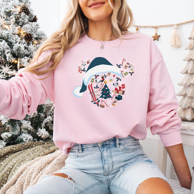 May include: A light pink sweatshirt featuring a Christmas-themed Mickey Mouse design. The design includes a Santa hat, a Christmas tree, and various holiday elements. The sweatshirt is a long-sleeved crew neck.