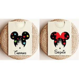 May include: Two white t-shirts featuring black silhouettes of Mickey Mouse and Minnie Mouse heads with a castle inside. The Mickey Mouse shirt has colourful hearts and the Minnie Mouse shirt has a red bow. The shirts are printed with the names "Thomas" and "Sophia".