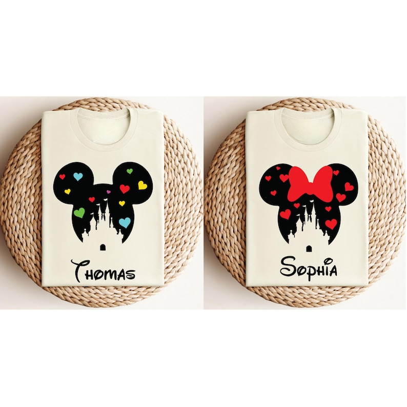 May include: Two white t-shirts featuring black silhouettes of Mickey Mouse and Minnie Mouse heads with a castle inside. The Mickey Mouse shirt has colourful hearts and the Minnie Mouse shirt has a red bow. The shirts are printed with the names "Thomas" and "Sophia".