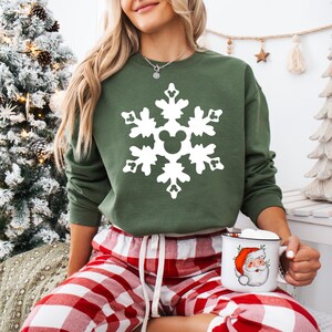 May include: Green sweatshirt with a white snowflake design featuring a Mickey Mouse head in the center. The person is wearing red and white plaid pajama pants and holding a mug with a Santa Claus graphic.