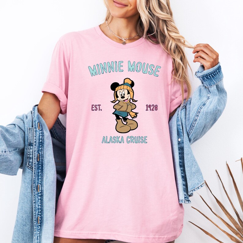 May include: Pink t-shirt featuring a Minnie Mouse graphic. The design includes the text "MINNIE MOUSE", "EST. 1928", and "ALASKA CRUISE". Minnie Mouse is depicted in winter attire, wearing a hat, scarf, and boots.