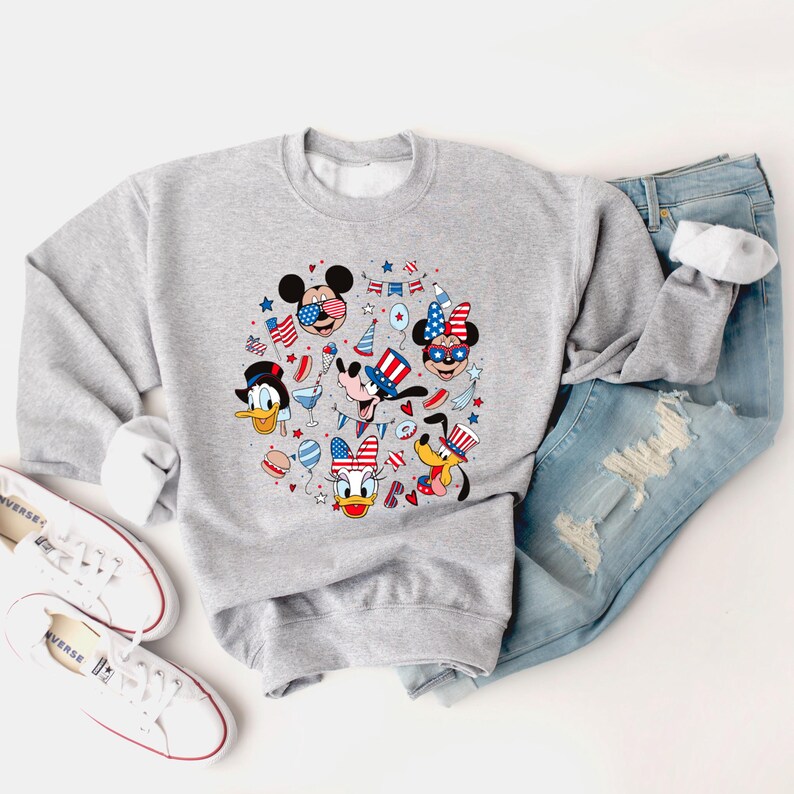 May include: A light gray sweatshirt featuring a circular graphic of cartoon characters in patriotic attire. The graphic includes Mickey Mouse, Minnie Mouse, Donald Duck, Daisy Duck, Goofy, and Pluto, with stars, stripes, and festive elements.