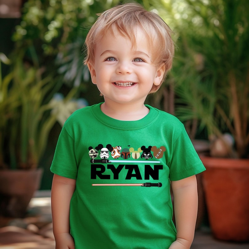 May include: A green t-shirt featuring a design with cartoon characters and the name "RYAN" in black. The design includes a lightsaber and various character icons. The shirt is short-sleeved and suitable for children.
