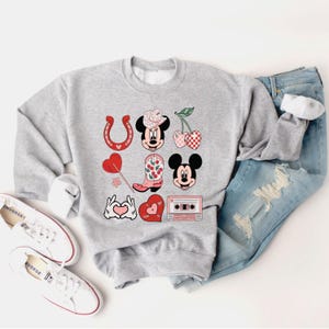 May include: A grey sweatshirt with a red, white and pink graphic design featuring Mickey Mouse, a horseshoe, a cowboy boot, cherries, a heart and a cassette tape.