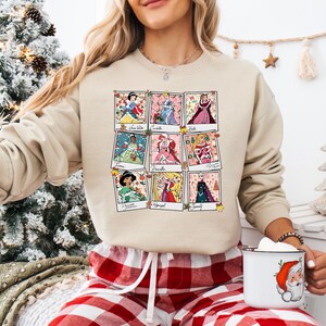 Puede incluir: Sudadera beige con una cuadr&iacute;cula de nueve retratos de princesas al estilo polaroid. Cada retrato muestra una princesa diferente con su nombre debajo. La sudadera se usa con pantalones de pijama a cuadros rojos y blancos.