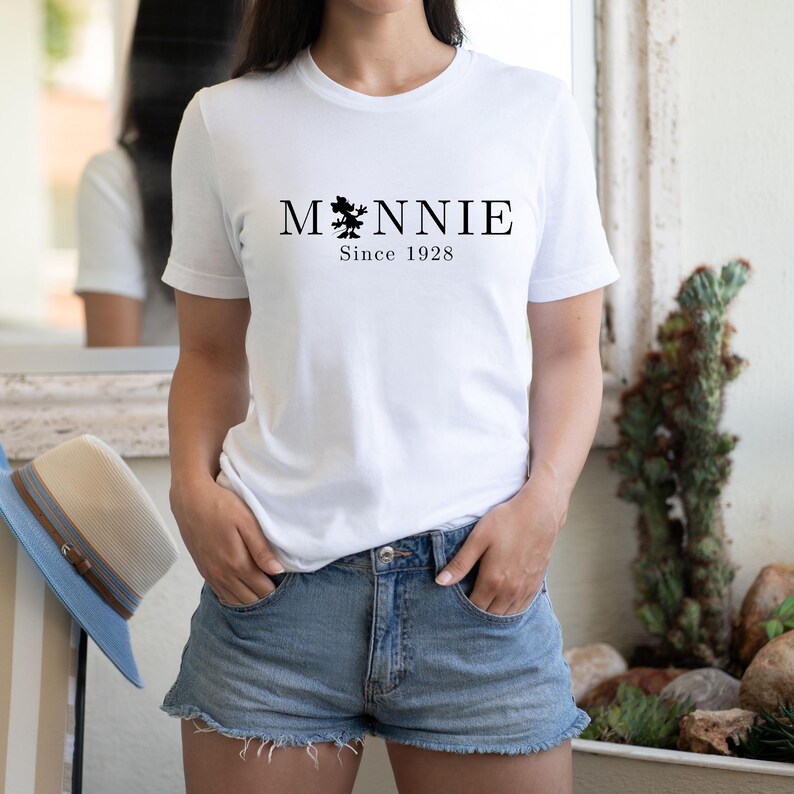 May include: White t-shirt with the word "Minnie" in black, featuring a silhouette of Minnie Mouse. Below the name is the text "Since 1928". The model is wearing denim shorts.