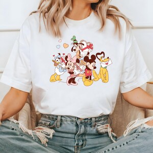 May include: White t-shirt with a graphic print of Mickey Mouse, Minnie Mouse, Donald Duck, Goofy, and Pluto. The characters are surrounded by pink hearts.