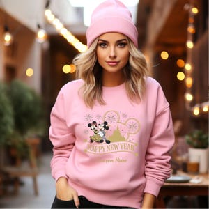 May include: Pink sweatshirt with a graphic of Mickey Mouse and Minnie Mouse in front of Cinderella's Castle with fireworks. The text "Happy New Year" and "Custom Name" are printed on the sweatshirt.
