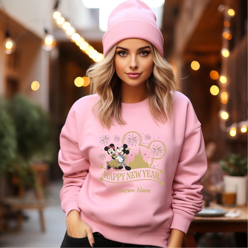 May include: Pink sweatshirt with a graphic of Mickey Mouse and Minnie Mouse in front of Cinderella's Castle with fireworks. The text "Happy New Year" and "Custom Name" are printed on the sweatshirt.
