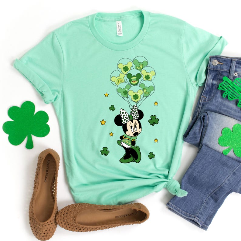 May include: A green t-shirt with a graphic of Mickey Mouse dressed as a leprechaun holding green balloons with shamrocks. The shirt is on a white background with a pair of brown shoes and blue jeans.