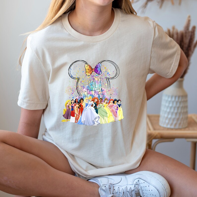 Puede incluir: Una camiseta blanca con un gr&aacute;fico de las orejas de Mickey Mouse con un lazo y el castillo de Disney en el centro. El castillo est&aacute; rodeado por un grupo de princesas Disney.