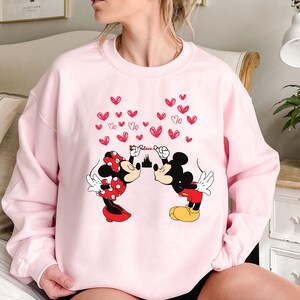 Puede incluir: Una sudadera rosa claro con un gr&aacute;fico de Mickey y Minnie Mouse bes&aacute;ndose, con corazones rojos y la palabra "Love" encima. Minnie lleva un lazo y una falda de lunares rojos. Mickey lleva pantalones cortos rojos y zapatos amarillos.