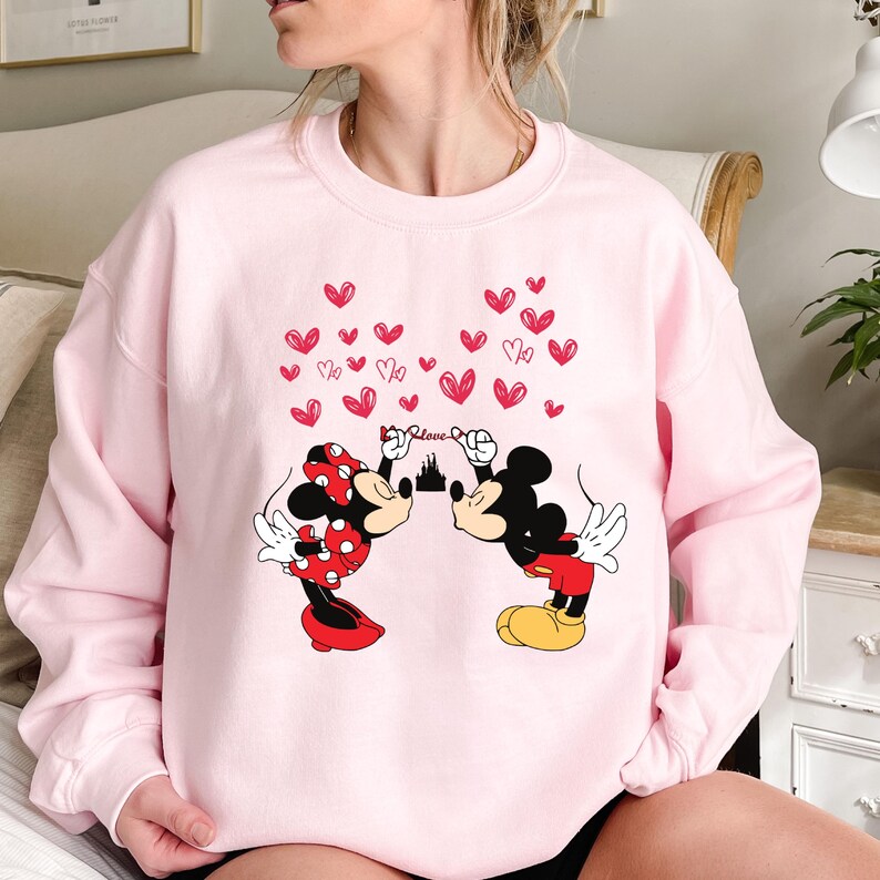 Puede incluir: Una sudadera rosa claro con un gr&aacute;fico de Mickey y Minnie Mouse bes&aacute;ndose, con corazones rojos y la palabra "Love" encima. Minnie lleva un lazo y una falda de lunares rojos. Mickey lleva pantalones cortos rojos y zapatos amarillos.