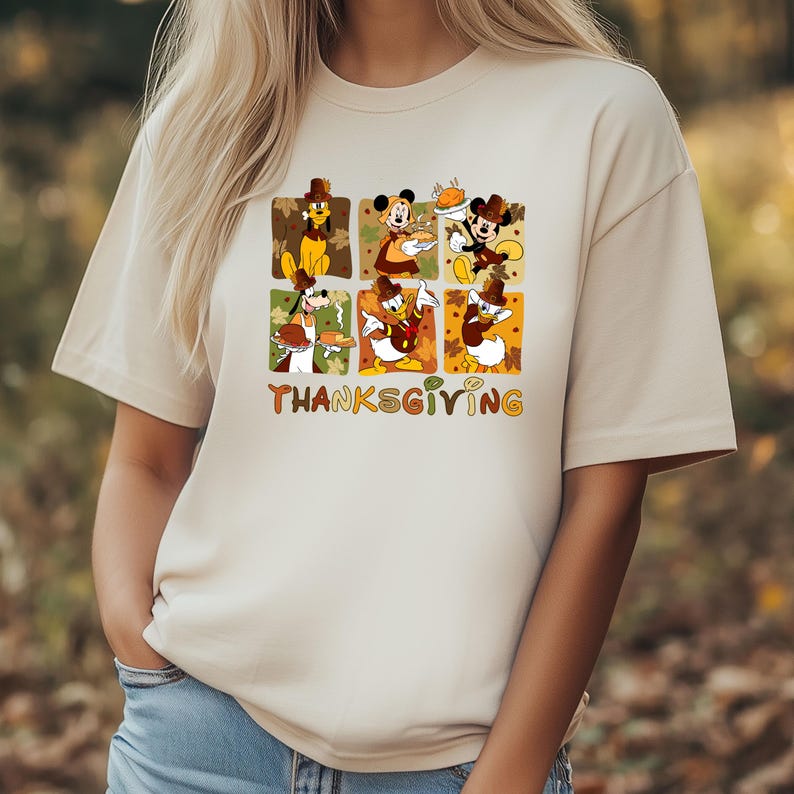 May include: Beige t-shirt featuring a Thanksgiving-themed graphic. The design includes cartoon characters like Mickey Mouse, Donald Duck, and Pluto, each in a Thanksgiving setting. The word "THANKSGIVING" is written in orange and yellow.
