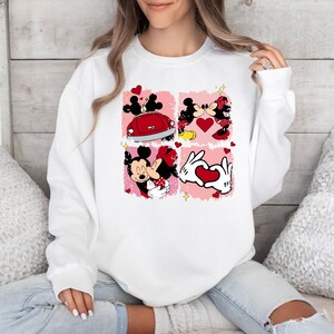 May include: White sweatshirt featuring a graphic print of Mickey Mouse and Minnie Mouse in four different scenes. The scenes include Mickey and Minnie kissing, Mickey and Minnie driving a red car, Mickey and Minnie hugging, and Mickey and Minnie making a heart shape with their hands.