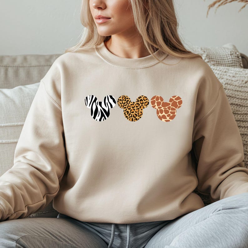 May include: Beige sweatshirt featuring three Mickey Mouse head silhouettes with animal print patterns: zebra, leopard, and giraffe. The sweatshirt has a crew neck and long sleeves, offering a casual and comfortable style.