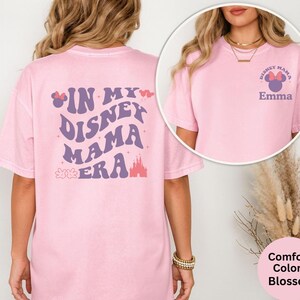 Puede incluir: Una camiseta rosa claro con el texto "IN MY DISNEY MAMA ERA" en morado y blanco. La parte posterior de la camiseta presenta un gr&aacute;fico de castillo. La parte delantera de la camiseta tiene un peque&ntilde;o gr&aacute;fico que dice "DISNEY MAMA" con un lazo de Minnie Mouse y el nombre "Emma".