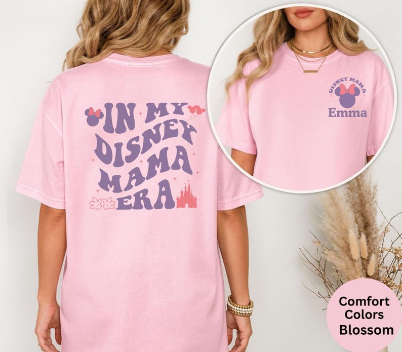 Puede incluir: Una camiseta rosa claro con el texto "IN MY DISNEY MAMA ERA" en morado y blanco. La parte posterior de la camiseta presenta un gr&aacute;fico de castillo. La parte delantera de la camiseta tiene un peque&ntilde;o gr&aacute;fico que dice "DISNEY MAMA" con un lazo de Minnie Mouse y el nombre "Emma".