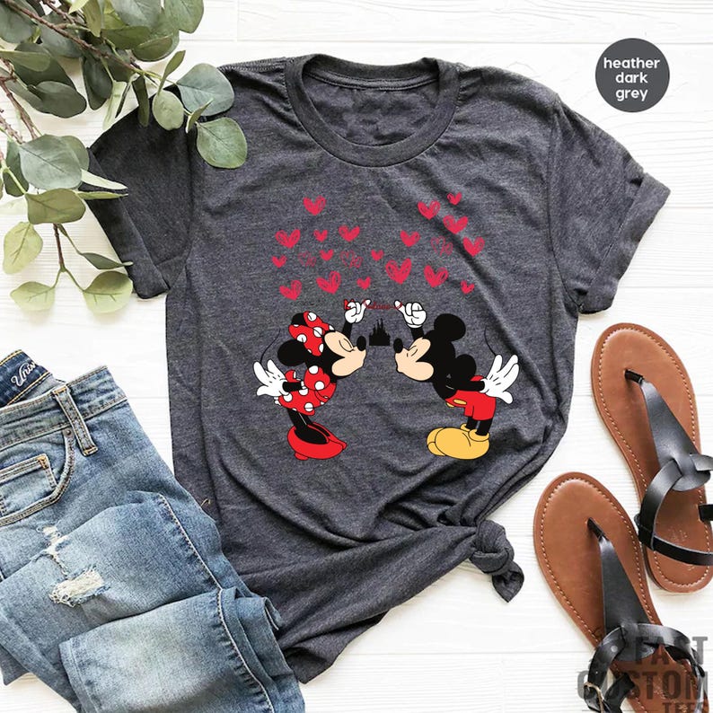 Puede incluir: Camiseta gris oscuro con Mickey y Minnie Mouse bes&aacute;ndose, gr&aacute;ficos de corazones rojos y una silueta de castillo. La camiseta se combina con vaqueros azules y sandalias marrones.