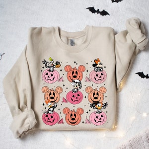 May include: A beige sweatshirt with a Halloween-themed design featuring Mickey Mouse, Minnie Mouse, Goofy, and Donald Duck dressed as skeletons inside pumpkins. The pumpkins are in shades of pink and orange.