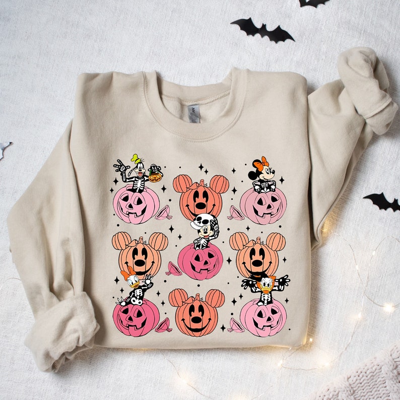 May include: A beige sweatshirt with a Halloween-themed design featuring Mickey Mouse, Minnie Mouse, Goofy, and Donald Duck dressed as skeletons inside pumpkins. The pumpkins are in shades of pink and orange.