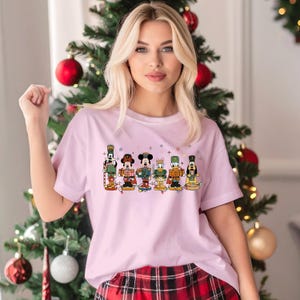 May include: A light pink t-shirt featuring a colorful graphic of Disney characters dressed as nutcrackers. The design includes Mickey Mouse, Donald Duck, and Goofy, with festive details. The shirt is a crew neck and has short sleeves.