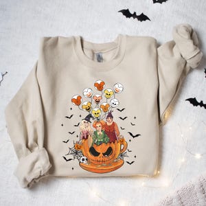 May include: A beige sweatshirt with a graphic print of three witches riding in a pumpkin-shaped teacup with Mickey Mouse balloons. The witches are wearing Halloween costumes and holding candy.