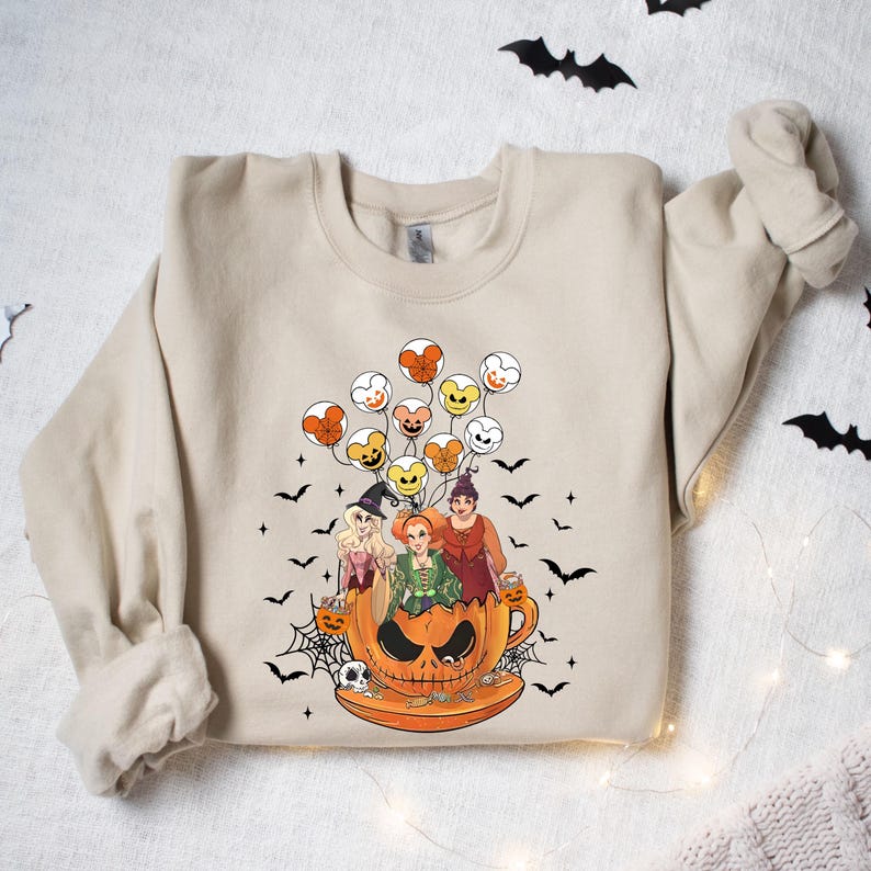 May include: A beige sweatshirt with a graphic print of three witches riding in a pumpkin-shaped teacup with Mickey Mouse balloons. The witches are wearing Halloween costumes and holding candy.