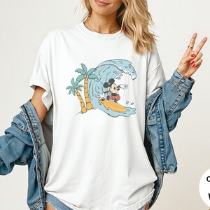 May include: White t-shirt featuring a cartoon Mickey Mouse surfing a wave. The design includes palm trees and a yellow surfboard. The shirt has the words "Comfort Colors White" on the bottom right.